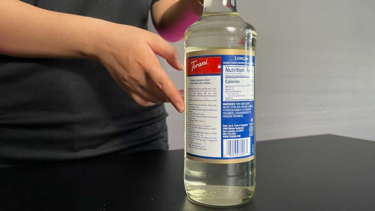 Ly siro Torani Nhãn trong suốt, ánh vàng nhẹ, đặt trên nền gỗ sáng